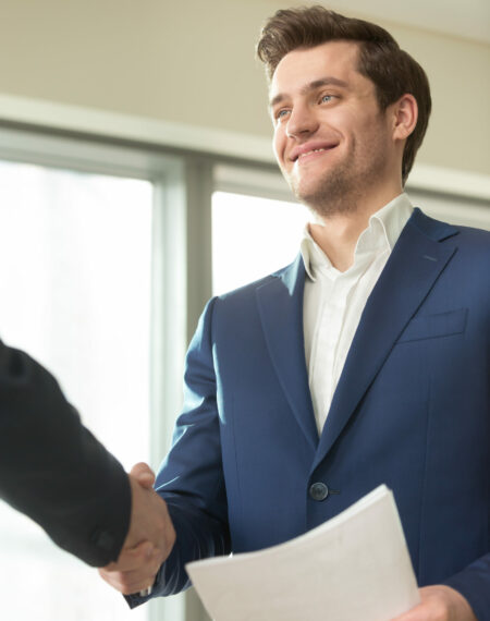 Confident smiling businessman with documents in hand handshaking with partner at business meeting in office. Presentable financial consultant welcoming client, congratulating investor with good deal